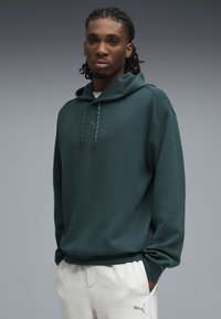 Felpa verde con cappuccio a sacco, cappuccio con coulisse e dettaglio logo discreto. Indossata con pantaloni della tuta grigi chiari, dotati di tasche laterali.