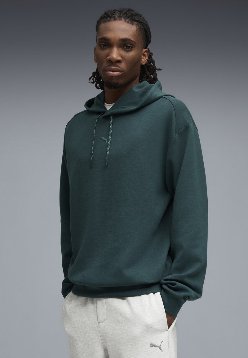 Sudadera verde con bolsillo canguro, capucha con cordón y detalle sutil del logo. Combinada con pantalones de chándal gris claro, que tienen bolsillos laterales.
