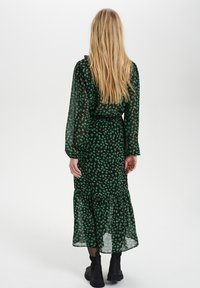 Lång svart blommig klänning med gröna mönster, genomskinligt tyg, långa ärmar och en lagerdelad nederkant. Bärs med svarta stövlar vid vristen.