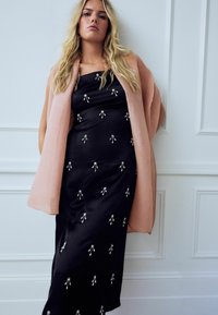 Robe en satin noir ornée d'embellissements floraux argentés, superposée avec un gilet en maille côtelée rose clair. La robe présente une silhouette élégante.
