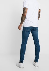 Hombre con camiseta blanca, jeans ajustados azules, zapatillas blancas, mostrando la espalda y con brazos tatuados contra un fondo neutro.