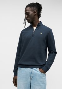 Μπλε ναυτικό φούτερ με φερμουάρ κατασκευασμένο από απαλό ύφασμα, με ψηλό γιακά και χρυσή λεπτομέρεια λογότυπου στο στήθος. Φοριέται με ανοιχτό μπλε τζιν.