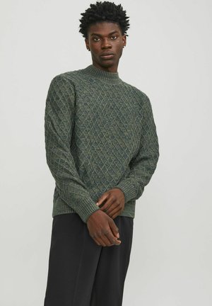 Sujeto con un suéter verde de punto que presenta un patrón de diamantes, puños acanalados y un cuello alto. Combina con pantalones negros.