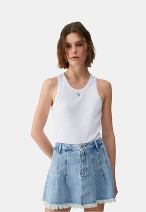 Mujer con camiseta sin mangas blanca de canalé y falda vaquera azul claro con dobladillo deshilachado, de pie frente a un fondo liso.