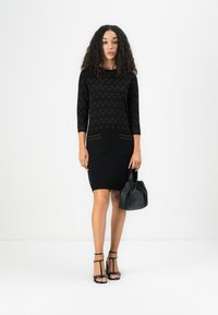 Robe noire à manches trois quarts, haut à motifs, jupe unie, poches avant, associée à un sac à main noir froncé et des talons à lanières.