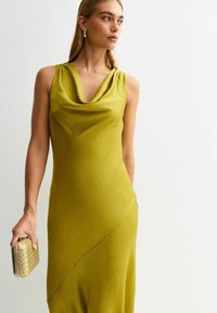 Klänning med cowl-halsringning och utan ärmar i olivgrön färg, med en mjuk, fallande textur och en asymmetrisk fåll. Modell som håller en texturerad kuvertväska.