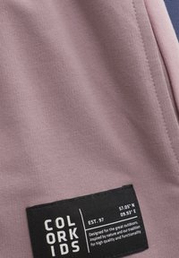 Ljus mauve tyg med en jämn textur och en liten svart etikett med vit text, inklusive varumärkesnamn och koordinater.