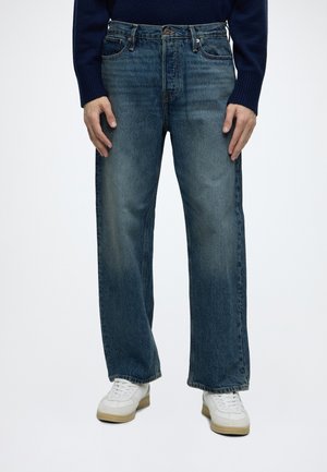 Personne portant un jean bleu ample avec un décoloration aux cuisses et des baskets blanches, debout les mains détendues le long du corps.