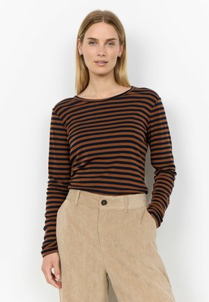Woman with straight blonde hair wearing a brown and black striped long-sleeve shirt and beige corduroy pants standing against a white background.