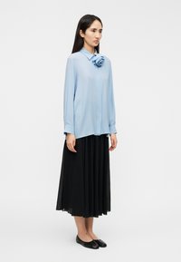 Μπλε μπλούζα με γιακά και διακοσμητικό λουλούδι, συνδυασμένη με μια μαύρη ρέουσα φούστα. Το μοντέλο στέκεται με μαύρα φλατ παπούτσια σε απλό φόντο.