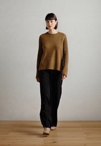 Bruine geribbelde gebreide trui met een ronde hals, gecombineerd met zwarte wijde broek en lichtbeige schoenen op een houten vloer.