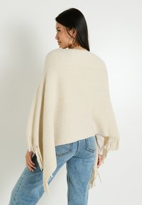 Poncho beige con flecos en el dobladillo, hecho de una tela texturizada. Estilizado sobre una blusa clara y combinado con jeans de tiro alto azules.