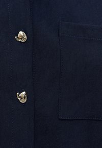 Tissu bleu marine foncé avec deux boutons dorés brillants de forme irrégulière à côté d'une poche rectangulaire cousue.