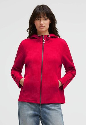 Woman wearing a textured red zip-up hoodie with hands in pockets and light blue jeans, standing against a plain light background.