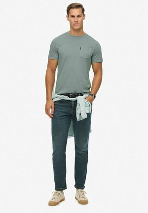 Grå kortærmet t-shirt med lomme, blå jeans og cremefarvede sneakers. Lysternet skjorte bundet om taljen, afslappet look.