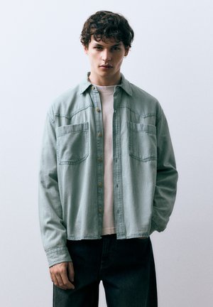 Chemise en velours côtelé vert clair avec ouverture boutonnée, deux poches poitrine et manches longues. Portée sur un t-shirt clair et un jean.