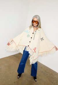 Γυναίκα με μακριά γκρίζα μαλλιά που φοράει υπερμεγέθη κρεμ πόντσο με κρόσσια και κεντημένα σύμβολα, μπλε τζιν, καφέ γυαλιά ηλίου και μαύρα μυτερά μποτάκια.