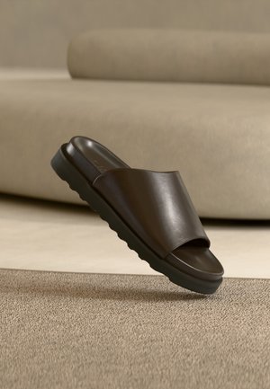 Dunkelbraune Lederslide-Sandale mit dicker Sohle auf beigem Teppich vor einem geschwungenen beigen Sofa.