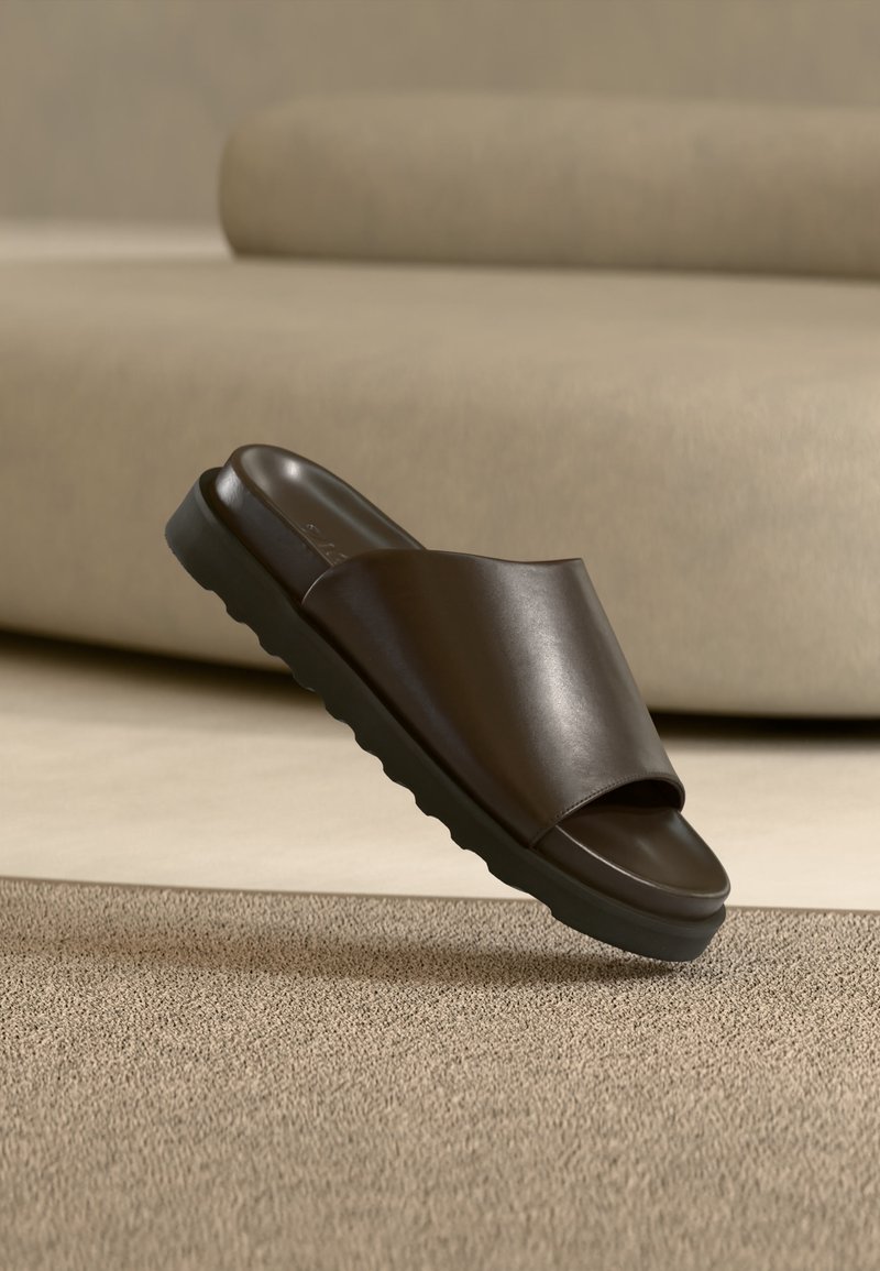 Sandale slide en cuir marron foncé avec une semelle épaisse sur un tapis beige devant un canapé beige courbé.