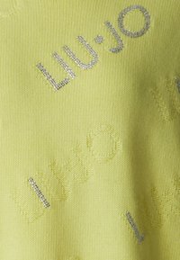 Sweater Liu Jo em malha amarelo suave, apresentando detalhes em relevo com o logo/textura e acentos de tachas prateadas. Textura suave com mangas curtas.