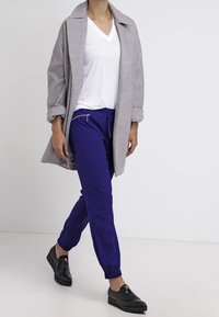 Hellgrauer übergroßer Mantel, weißes V-Ausschnitt-T-Shirt, lila schmal geschnittene Hose und schwarze Slipper mit dekorativen Reißverschlüssen.