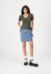 Top verde oliva a maniche corte con centro arricciato, abbinato a una gonna di jeans azzurro chiaro con orli grezzi e tre cuciture verticali. Scarpe nere con suola spessa.