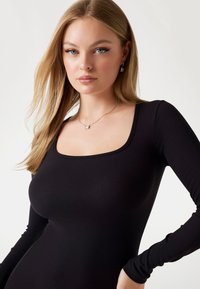 Rochie lungă neagră cu mânecă lungă, de tip côté, cu decolteu pătrat și siluetă ajustată. Modelul poartă un colier cu pandantiv argintiu și cercei.