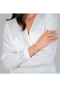 Pulsera de plata con cuentas de perlas y un charm de concha dorada, llevada en una muñeca, con una blusa blanca parcialmente visible en el fondo.