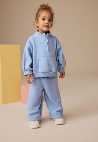 Lyseblå sweatshirt med halv lynlås og frontlomme, som er sat sammen med matchende løstsiddende sweatpants. Båret med hvide sneakers.