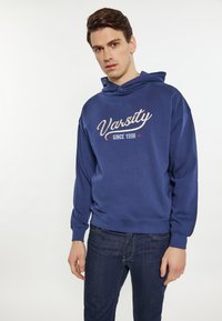 Sötétkék kapucnis pulóver, fehér és piros "Varsity" logóval és "Since 1998" részlettel. Puha tapintású, laza stílusú.
