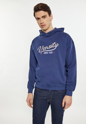Námořnická modrá mikina s kapucí, která má bílý a červený nápis "Varsity" a detail "Since 1998". Měkká textura, volnočasový design.