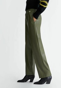 Pantalones verde oliva de tiro alto, de material sintético y con un corte relajado, que cuentan con bolsillos laterales y un detalle en dorado en la cintura. Botas negras de tacón.