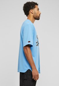 Šviesiai mėlynas trumpomis rankovėmis marškinėliai su tekstūruotu audiniu, kurių priekyje yra ryški grafika, o rankovėje yra mažas etiketės akcentas.