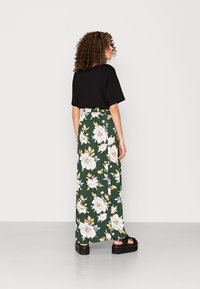 T-shirt noir oversize associé à une longue jupe florale verte ornée de fleurs blanches et jaunes, portée avec des sandales à plateforme noires.