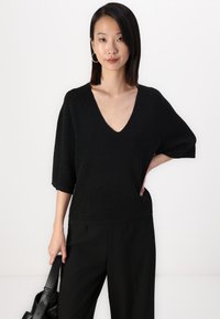 Top preto de malha com decote em V profundo, mangas curtas e largas, e um acabamento texturizado. Combinado com calças pretas de cintura alta e uma mala preta.