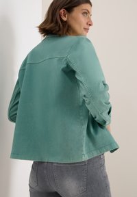 Femme avec les cheveux bruns courts portant un haut à manches longues teal et un jean noir délavé, tournée sur le côté devant un fond clair et uni.