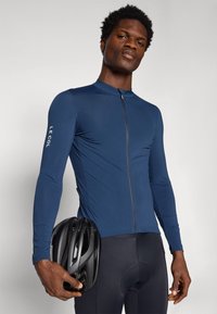 Maillot de cyclisme à manches longues bleu marine avec une fermeture éclair à l'avant et un logo sur la manche. Cuissard de cyclisme rembourré noir, tenant un casque noir.