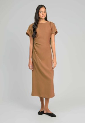 Une femme porte une robe midi marron à manches courtes avec des détails à boutons sur une épaule, associée à des chaussures noires sans lacets.
