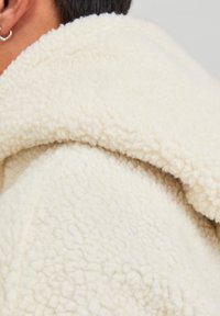 Giacca in pile color crema con superficie testurizzata e soffice e cappuccio arrotondato. Il materiale appare morbido e accogliente.