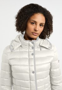 Witte gewatteerde jas met een hoge kraag, glanzende textuur en knoopaccenten; heeft trekkoorden en een quilted ontwerp.