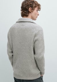 Giovane uomo con capelli castani ricci indossa un maglione lavorato a maglia grigio chiaro e pantaloni scuri, di spalle, su uno sfondo grigio chiaro uniforme.