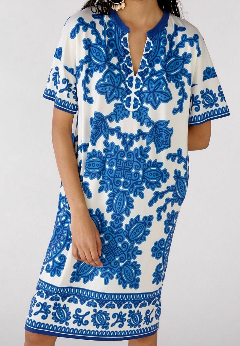 Femme portant une robe blanche mi-longue à manches courtes avec des motifs floraux et feuillages bleus complexes.