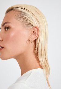 Boucle d'oreille en or portée à l'oreille gauche, avec des cheveux blonds lisses et raides. La peau présente un léger éclat, avec une chemise blanche texturée.