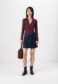 Burgundska rebrasta dolga majica, temno modra denim A-linijska krila, črne gležnjarje in rjava torba preko ramena, postavljene proti enostavni ozadju.