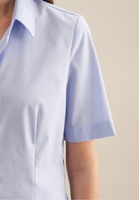 Ljusblå skjorta med kort ärm och spetsig krage. Slät textur och skräddarsydd passform, med subtila sömmar längs kroppen för att ge form.