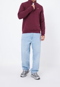 Tmavě červený pulovr s částečným zipem a logem na hrudi, v kombinaci s volnými světle modrými džíny a šedými teniskami s černými detaily.