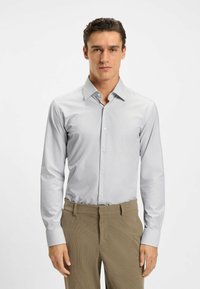 Camicia da uomo a maniche lunghe in grigio chiaro con sottili righe bianche, collo button-down e polsini a contrasto, abbinata a pantaloni color sabbia.