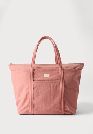 Grand sac fourre-tout en tissu rose doux avec deux poignées et une poche avant, sur un fond clair uni.