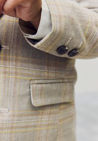 Blazer beige à carreaux avec tissu texturé comportant des lignes jaunes et orange, deux boutons foncés, et une petite poche sur le côté gauche.