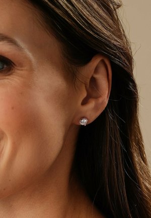 Nahaufnahme eines Frauenohres mit einem kleinen runden Ohrstecker aus klarem Edelstein, umrahmt von dunkelbraunem Haar, das hinter das Ohr gesteckt ist.